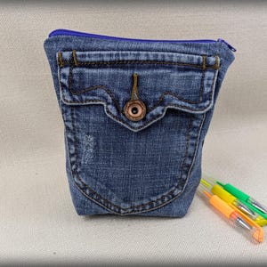 May include: A blue denim zippered pouch with a large front pocket and a brown button. The pouch is made from recycled denim and is perfect for carrying small items.