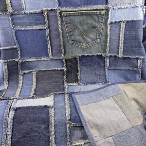 May include: A patchwork quilt made from various shades of blue denim. The quilt features a variety of denim squares and rectangles, with a denim pocket sewn onto the surface. The edges of the denim pieces are frayed, adding texture.