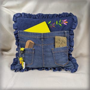 Puede incluir: Un cojín cuadrado de mezclilla con borde de volantes y detalle floral bordado. El frente tiene dos bolsillos con un peine de madera y un cuaderno beige, con un bloc de notas amarillo detrás. Un llavero de pato amarillo cuelga de un bolsillo.