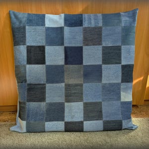 Puede incluir: Un cojín cuadrado con un diseño de patchwork hecho de varios tonos de mezclilla azul. La superficie del cojín está dividida en una cuadrícula de cuadrados, creando un patrón texturizado y visualmente atractivo. El cojín está sobre una alfombra.