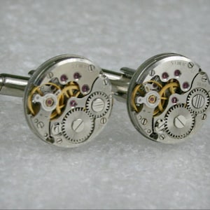 May include: A pair of silver cufflinks with a circular design featuring exposed watch gears and small pink jewels.