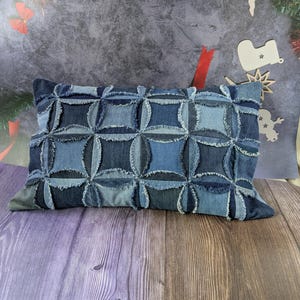 Puede incluir: Un cojín rectangular de mezclilla con un diseño de patchwork. El cojín presenta un patrón de cuadrados y círculos en varios tonos de mezclilla azul. Los bordes de las piezas de mezclilla están deshilachados, agregando textura.
