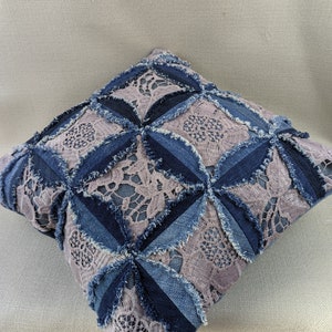 Puede incluir: Una almohada cuadrada con un diseño de patchwork hecho de mezclilla y encaje. La almohada es azul y gris con un borde deshilachado.