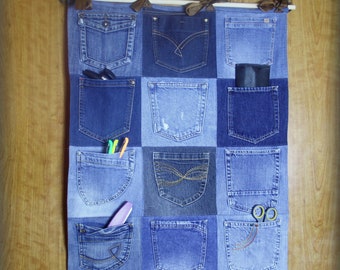 Op de afbeelding: Een wandorganizer gemaakt van denim met 12 zakken in verschillende maten en vormen. De organizer hangt aan een houten muur en heeft bruine lintjes.
