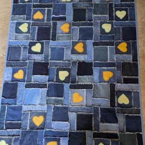 Puede incluir: Una colcha de retazos hecha de tela vaquera en una variedad de tonos azules. La colcha está decorada con corazones amarillos y azul claro.