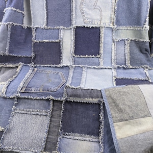 Può includere: Una trapunta patchwork realizzata con denim in varie tonalità di blu. La trapunta presenta quadrati e rettangoli di diversi lavaggi di denim, tra cui blu chiaro, medio e scuro. Alcuni pezzi includono tasche. I bordi dei pezzi di denim sono sfilacciati.