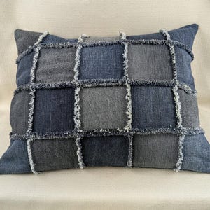 Puede incluir: Una almohada rectangular con un diseño de patchwork hecho de diferentes tonos de mezclilla azul. La almohada tiene un borde deshilachado y está hecha de materiales reciclados.