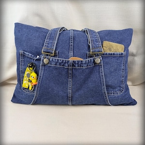 May include: A blue denim tote bag constructed from repurposed overalls, featuring two shoulder straps with metal buckles, multiple exterior pockets, a yellow duck keychain, and a small tan wallet tucked into one of the pockets.