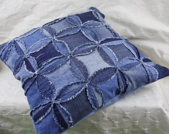 May include: A blue denim pillow with a patchwork design featuring a repeating flower pattern. The pillow is made from different shades of blue denim and has a frayed edge.