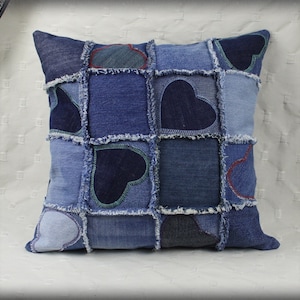 Puede incluir: Una almohada de mezclilla azul con un diseño de patchwork que presenta múltiples formas de corazón. La almohada está hecha de mezclilla reciclada y tiene un borde deshilachado.