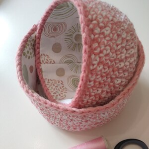 May include: Two pink crocheted baskets with a white and brown patterned fabric lining. A spool of pink thread and a pair of silver scissors are on a white surface.