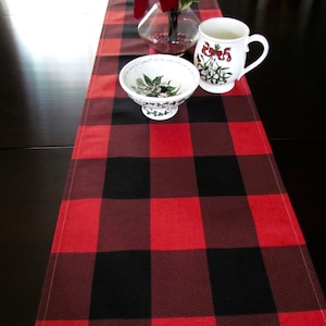 May include: Red and black buffalo plaid table runner with a white ceramic mug and bowl with a floral design on a black table.