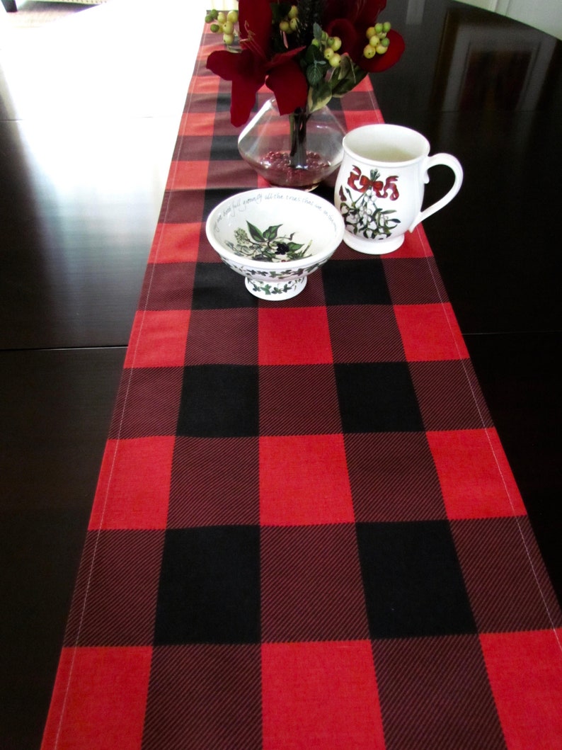 May include: Red and black buffalo plaid table runner with a white ceramic mug and bowl with a floral design on a black table.
