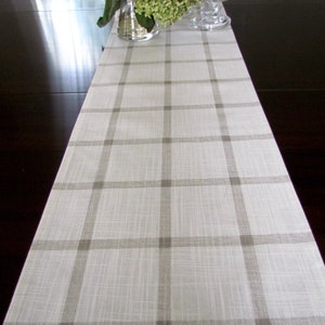 May include: A white table runner with a brown plaid pattern. The runner is on a dark wood table with a vase of green flowers and candles.