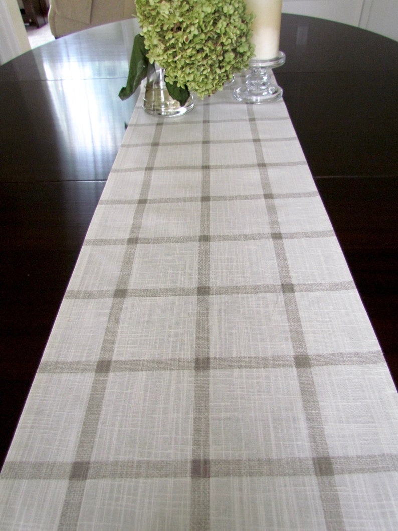 May include: A white table runner with a brown plaid pattern. The runner is on a dark wood table with a vase of green flowers and candles.