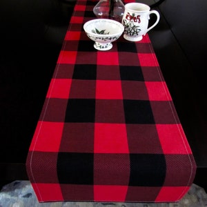 May include: Red and black buffalo plaid table runner with a white stitched edge. The runner is on a dark brown table with a white bowl and a white mug with a floral design.