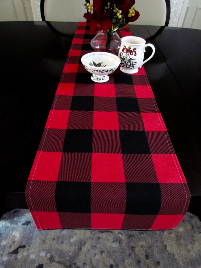 May include: Red and black buffalo plaid table runner with a white stitched edge. The runner is on a dark brown table with a white bowl and a white mug with a floral design.