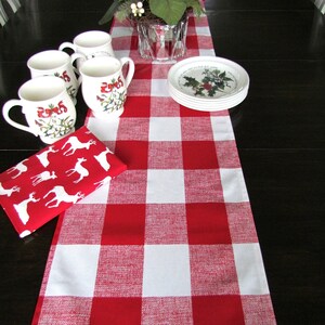 May include: A red and white checkered table runner with a white background and red squares. The runner is on a dark wood table with four white mugs with a red and green holiday design and a plate with a holiday design.