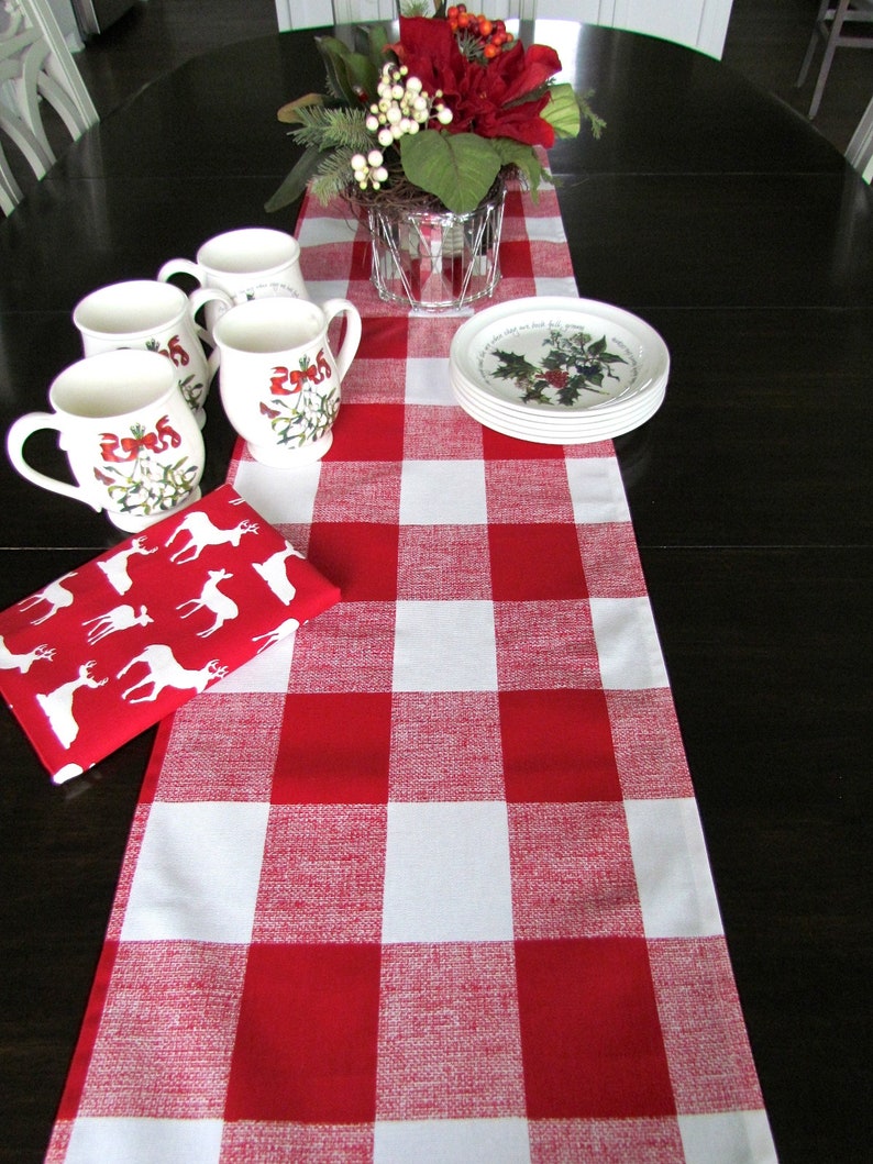May include: A red and white checkered table runner with a white background and red squares. The runner is on a dark wood table with four white mugs with a red and green holiday design and a plate with a holiday design.