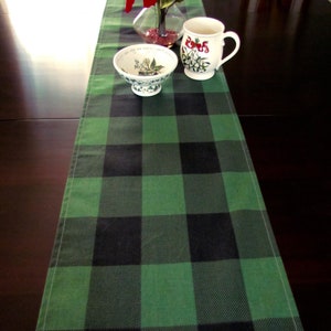 May include: A green and black buffalo plaid table runner with a white ceramic bowl and a white ceramic mug with a floral design on a dark brown wooden table.