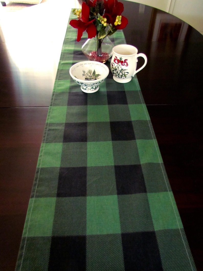 May include: A green and black buffalo plaid table runner with a white ceramic bowl and a white ceramic mug with a floral design on a dark brown wooden table.