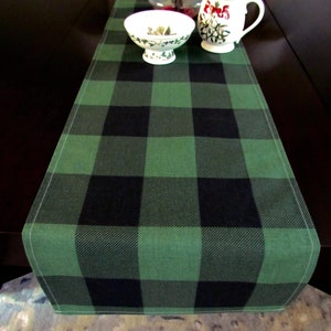 May include: A green and black buffalo plaid table runner. The runner is made of a soft, woven fabric and has a subtle sheen.