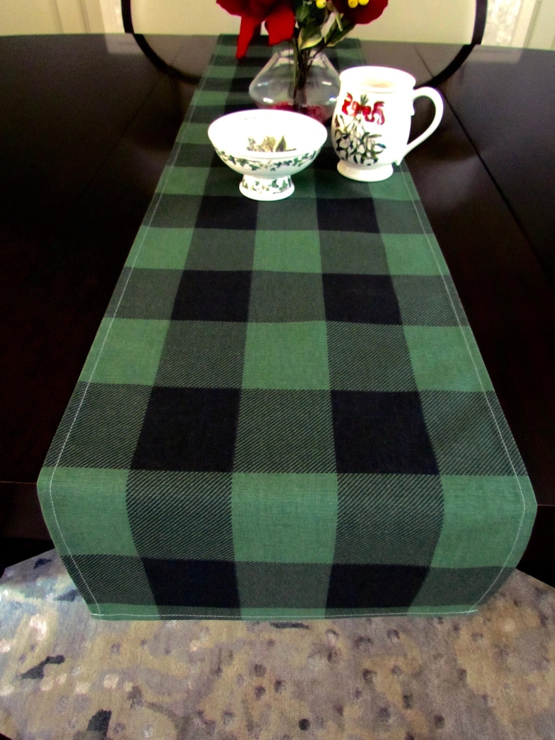 May include: A green and black buffalo plaid table runner. The runner is made of a soft, woven fabric and has a subtle sheen.