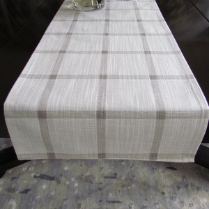 May include: A white and brown plaid table runner with a subtle textured weave. The runner is laid out on a dark brown table with a gray and white patterned rug underneath.