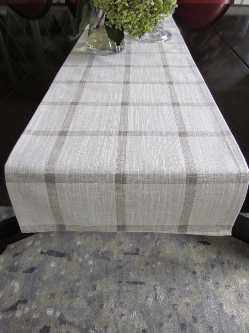 May include: A white and brown plaid table runner with a subtle textured weave. The runner is laid out on a dark brown table with a gray and white patterned rug underneath.