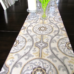May include: A decorative table runner with a repeating pattern of gray, beige, and brown circular and geometric shapes. The runner is placed on a dark wood table, with glass candle holders and a vase of purple tulips.