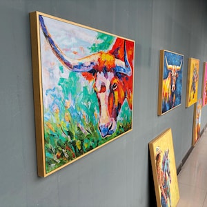 May include: A series of framed abstract paintings featuring a longhorn steer. The lead painting is a close-up of the steer's head, with vibrant colors of red, orange, blue, and green. The paintings are displayed on a gray wall.