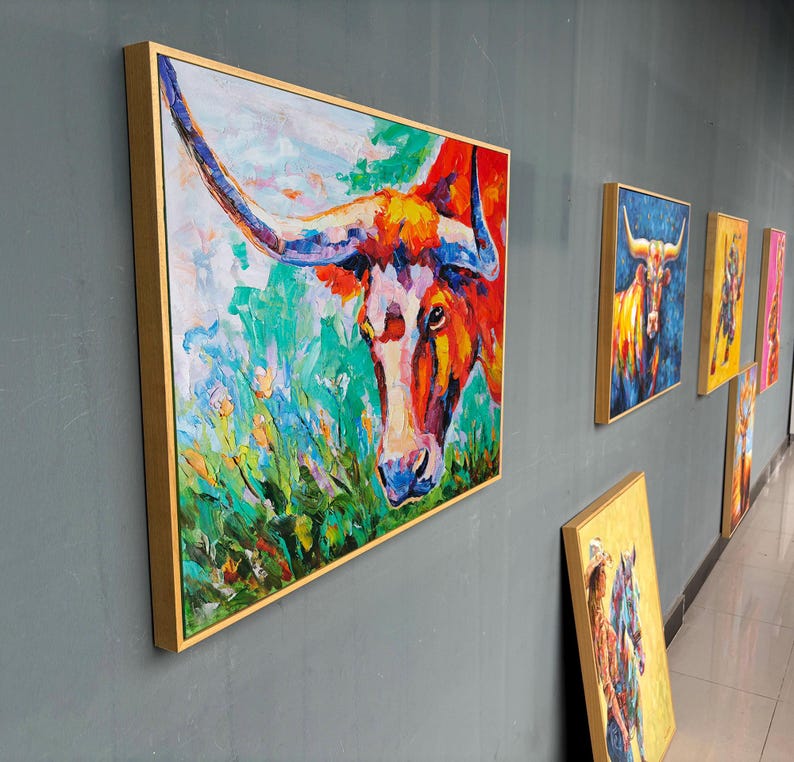 May include: A series of framed abstract paintings featuring a longhorn steer. The lead painting is a close-up of the steer's head, with vibrant colors of red, orange, blue, and green. The paintings are displayed on a gray wall.