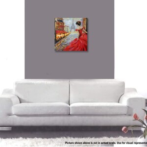 May include: An oil painting depicts a woman in a red dress walking on a cobblestone street in Paris. The Eiffel Tower is visible in the background. The artwork is displayed on a gray wall above a white sofa.