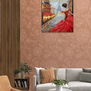 May include: A framed painting depicts a woman in a red dress walking down a Parisian street, with the Eiffel Tower in the background. The artwork is displayed on a floral patterned wall, above a sofa and chair. The room has a modern design.