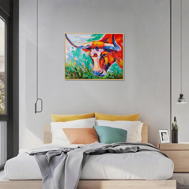 May include: Colorful abstract painting of a longhorn bull. The artwork features a vibrant palette of red, orange, and blue hues, with a focus on the bull's face and horns. The painting is framed and displayed above a bed with colorful pillows.