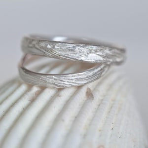 Puede incluir: Dos anillos de plata con un diseño texturizado de madera, apilados uno encima del otro sobre una concha blanca.