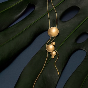 May include: A gold necklace with a long chain and multiple round pendant charms. The necklace is displayed on a dark green leaf.