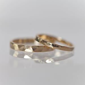 Könnte beinhalten: Zwei goldene, gehämmerte Trauringe, einer etwas größer als der andere, liegen übereinander. Die Ringe sind glatt und glänzend, mit einer leicht strukturierten Oberfläche.