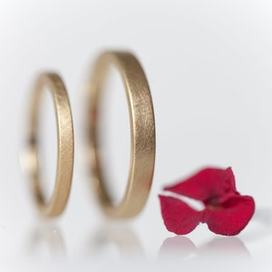 May include: Two gold wedding bands with a brushed finish, lying on a white surface next to a single red flower petal.