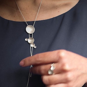 May include: A silver necklace with a long chain and multiple round silver pendants. The necklace is worn by a person with a dark blue shirt.