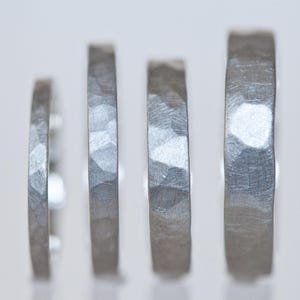 Może przedstawiać: Cztery srebrne pierścionki o młotkowanej fakturze są prezentowane na białym tle. Pierścionki r&oacute;żnią się szerokością, prezentując teksturowane, metaliczne wykończenie. Pierścionki są prawdopodobnie na ślub lub zaręczyny.