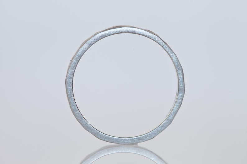 Może przedstawiać: Srebrny pierścionek o teksturowanym, młotkowanym wyglądzie. Pierścionek jest okrągły i wydaje się być wykonany z metalu. Design jest prosty i elegancki, odpowiedni do codziennego noszenia. Obręcz pierścionka ma szerokość około 0,2 cm.