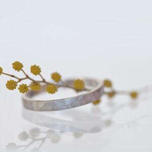 Może przedstawiać: Srebrny pierścionek o młotkowanej fakturze przeplatany jest gałązką ż&oacute;łtych kwiat&oacute;w. Pierścionek odbija się na białej powierzchni, tworząc efekt lustra. Obraz to zbliżenie, podkreślające szczeg&oacute;ły biżuterii i naturalnych element&oacute;w.