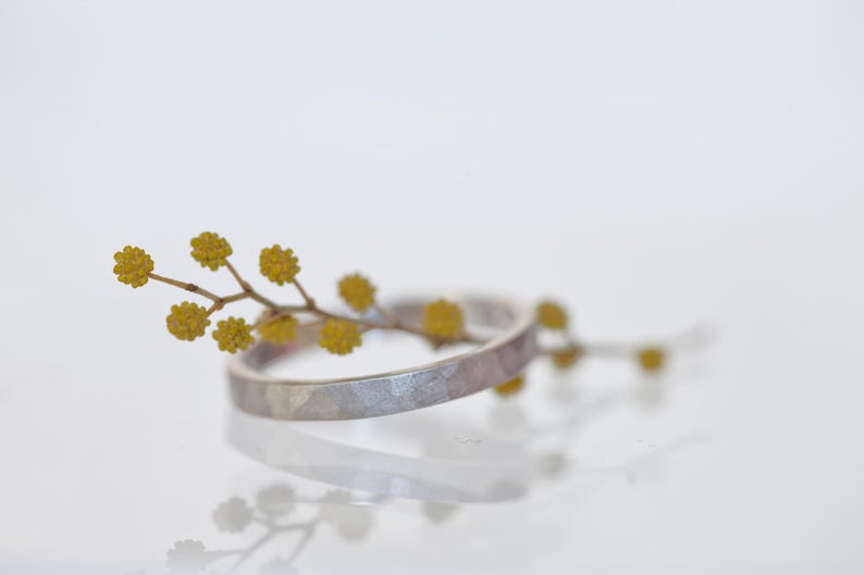 Może przedstawiać: Srebrny pierścionek o młotkowanej fakturze przeplatany jest gałązką ż&oacute;łtych kwiat&oacute;w. Pierścionek odbija się na białej powierzchni, tworząc efekt lustra. Obraz to zbliżenie, podkreślające szczeg&oacute;ły biżuterii i naturalnych element&oacute;w.