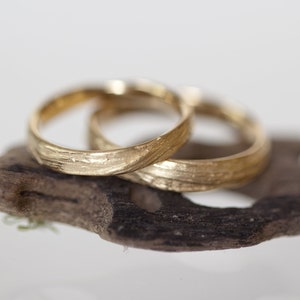 May include: Two gold wedding bands with a textured, wood-grain design. The bands are intertwined and resting on a piece of driftwood.