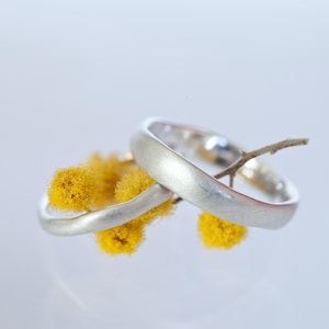Op de afbeelding: Twee zilveren trouwringen met een geborstelde afwerking, verweven met gele mimosa bloemen.