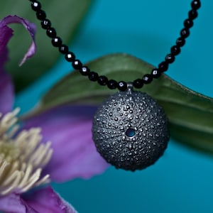 May include: A silver sea urchin pendant hangs from a black beaded necklace. The pendant is detailed with a textured surface and a small hole in the center. The necklace is displayed against a blue background with a purple flower.