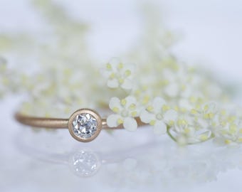 May include: A delicate gold ring with a bezel-set, round, clear gemstone. The ring is set against a soft, blurred background of white flowers. The ring's reflection is visible on the surface.