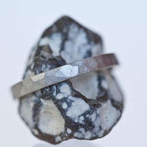 Może przedstawiać: Srebrny pierścionek o młotkowanej fakturze spoczywa na nakrapianym szaro-białym kamieniu. Pierścionek ma płaską, pasmową konstrukcję. Kamień ma szorstki, naturalny wygląd, kontrastujący z metalicznym połyskiem pierścionka.