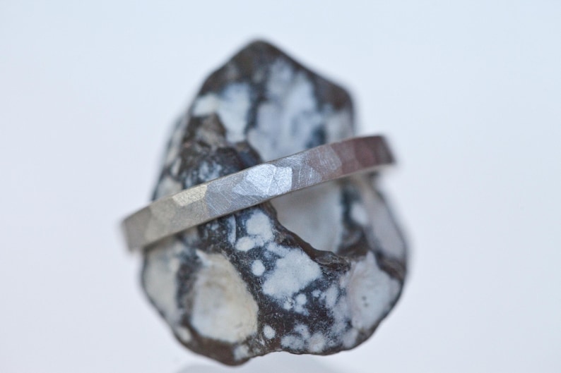 Może przedstawiać: Srebrny pierścionek o młotkowanej fakturze spoczywa na nakrapianym szaro-białym kamieniu. Pierścionek ma płaską, pasmową konstrukcję. Kamień ma szorstki, naturalny wygląd, kontrastujący z metalicznym połyskiem pierścionka.